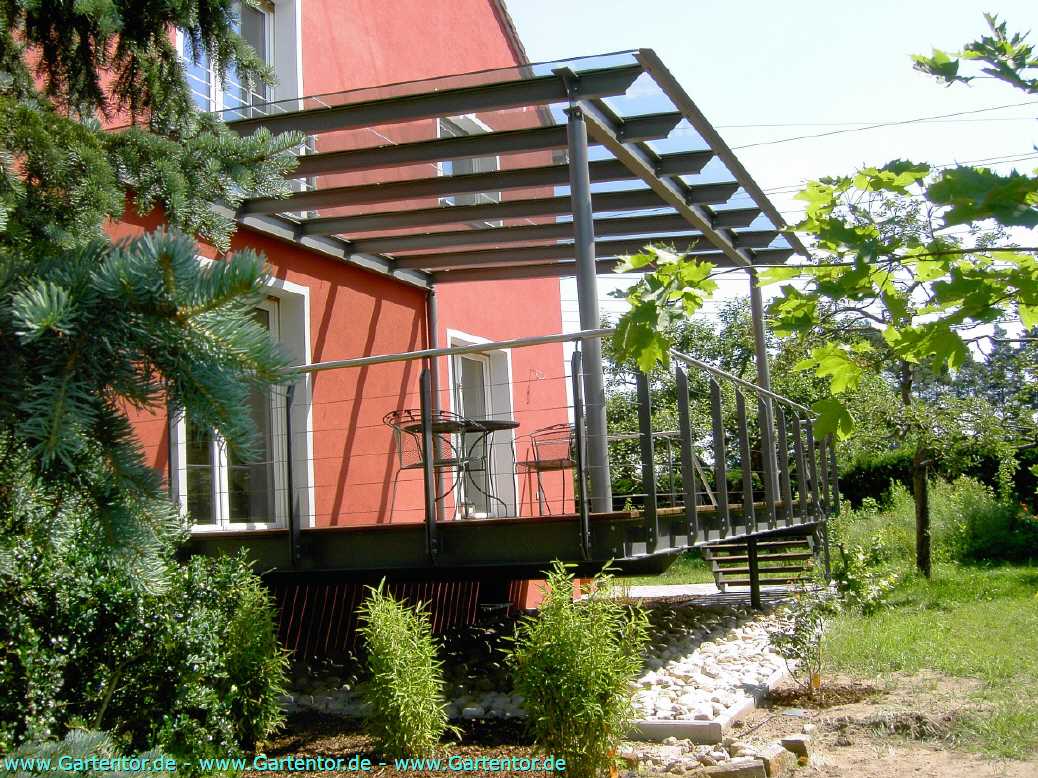 Erh&ouml;hte Terrasse mit moderner Terrassen&uuml;berdachung aus Metall und Glasdach an einem roten Wohnhaus, mit Edelstahlgel&auml;nder, Treppe in den Garten und Sitzgruppe.